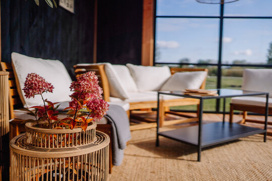 Close-up of a cozy patio with pink hydrangeas in a rattan planter, white cushioned chairs, a black metal table, and large windows revealing a scenic view.