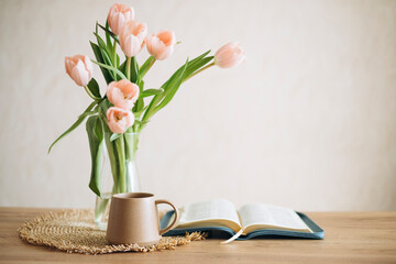 Open Bible on a wooden table and ceramic coffee mug and a vase with tulips.