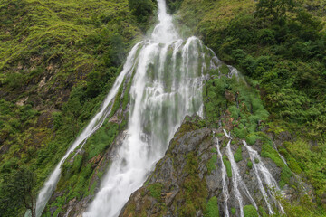 A stunning waterfall cascades down rocky cliffs in the mountains, surrounded by lush greenery, creating a peaceful and refreshing scene of untouched natural beauty.