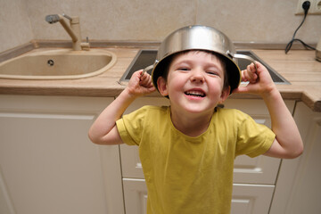 Happy boy wearing a pot on his head like a helmet in the kitchen. Child playing and having fun at...