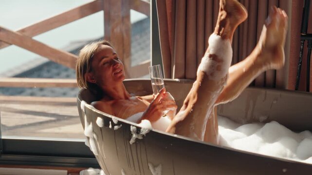 Happy young woman enjoys a hot bubble bath while sipping champagne in a cozy wooden cabin. Morning sunlight fills the room, creating a serene atmosphere of luxury, relaxation, and self-care. Weekend