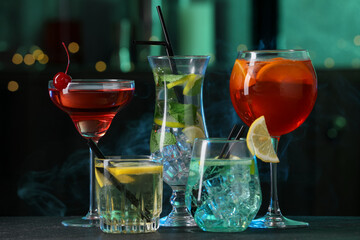 Glasses of tasty cocktails on dark table against blurred background with smoke, bokeh effect
