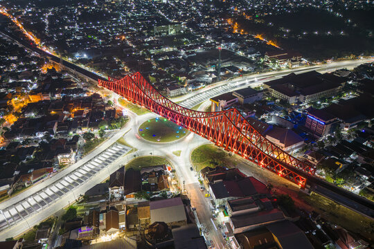 Aerial view of a vibrant red bridge arches gracefully over a bustling roundabout, amidst the twinkling cityscape at night, Surakarta, Central Java, Indonesia.