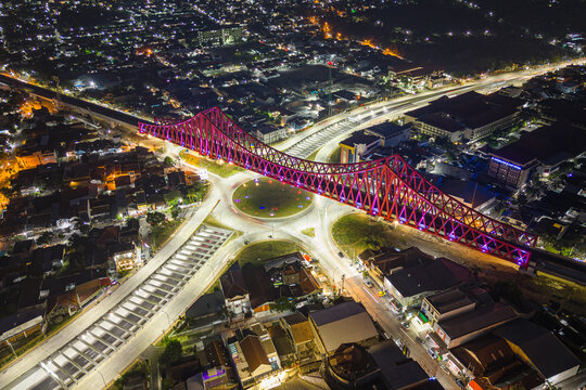 Aerial view of a vibrant red bridge illuminated against the dark sky and cityscape, connecting bustling streets and illuminated roundabouts, Surakarta, Central Java, Indonesia.
