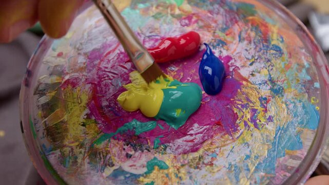 Close-up of an artist's brush dipping into pink and yellow paint on a palette.