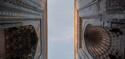 Looking upward between two ornate mausoleum portals in Samarkand, Uzbekistan, with intricate Persian-style muqarnas vaulting and turquoise mosaic tiles highlighted by golden sunlight