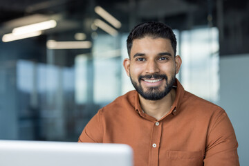 Young indian businessman confidently smiling, wearing a brown shirt, and sitting in a contemporary office setting with a laptop, conveying professionalism and success