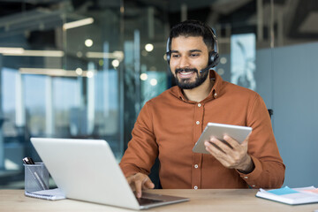 Smiling indian man wearing a headset and using a laptop and digital tablet, providing excellent customer service operating in a modern office or remote work setting
