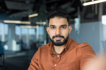 Young indian man in brown shirt confidently taking a selfie in a modern office, creating...