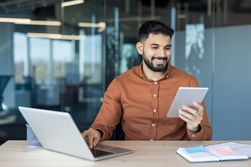 Young businessman happily working at office desk, holding a digital tablet while typing on a laptop, demonstrating efficiency, connectivity, and modern technology integration in daily tasks