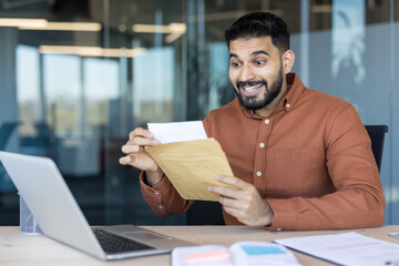 Fototapeta premium Excited young professional man sitting at his office desk opening a brown envelope with a surprised happy expression, learning about a promotion or financial success