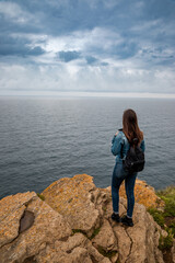 Contemplative traveler gazes at the vast ocean from dramatic rocky cliff on overcast day, seeking inspiration and adventure on a solo journey along the scenic coastline