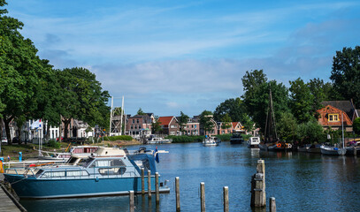 Canal in the village of Blokzijl, Overijssel, lined with moored boats, traditional houses, and a white drawbridge under a clear summer sky, surrounded by green trees and peaceful residential buildings