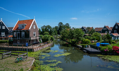 Obraz premium Traditional wooden houses with red roofs line a calm canal in Marken, North Holland. The foreground features lily pads on the water, a parked bicycle, and gardens with blooming flowers.