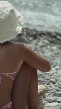 Beach woman hat, sitting on pebble beach, looking at sparkling sea. Vertical video