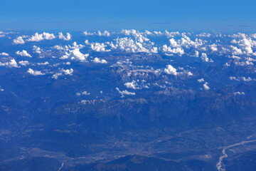 Alps as seen from an airplane with view of snow capped mountains peaking through layer of fluffy clouds under blue sky. Mountain range is major European tourist destination in Italy and Switzerland