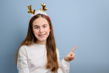 Smiling girl with sequin reindeer antlers pointing up on blue background. Festive portrait for Christmas and winter celebrations.