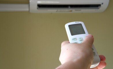 Hand holds air conditioner remote control with display showing 20 degrees Celsius temperature, blurred aircon on wall, comfortable temperature on split system. Heat and home interior equipment