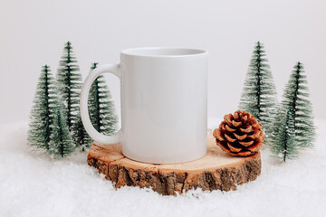 White mockup mug on wooden podium in the snow with christmas tree cone and christmas trees on white background. Ceramic mug with copy space for winter design, christmas and new year gift, print.