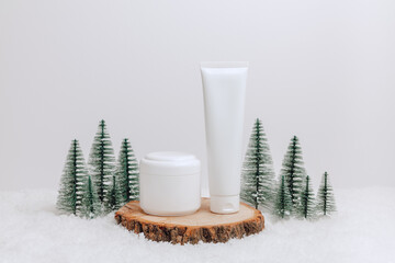 Tube with facial cream and bottle of body lotion on wooden podium in the snow with christmas trees at the background. Winter skincare mockup containers