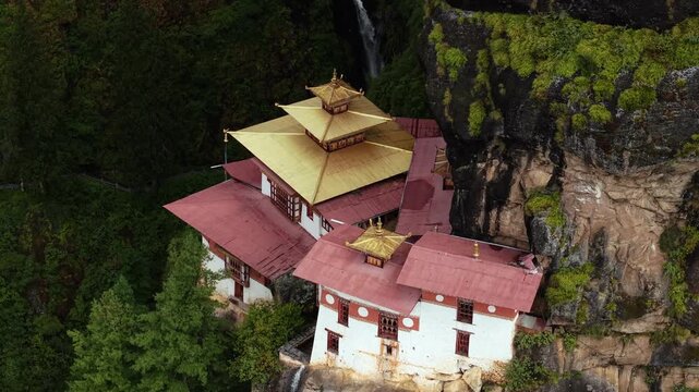 Aerial view of the Tigers Nest Temple clinging to the cliffside, a cascade of white water flowing beside it, amid lush greenery, Paro, Bhutan.