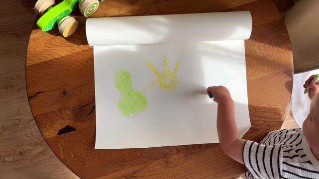 A small child draws with crayons on paper, view from above. A child's first drawing, development through painting, and home art.