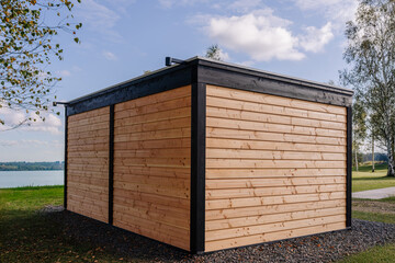 Modern wooden building with natural timber siding and black trim, located by a scenic lake, surrounded by greenery and gravel ground.