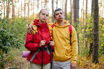 A couple stands in a forest looking unhappy while holding hiking gear. They are dressed for autumn weather in jackets and backpacks