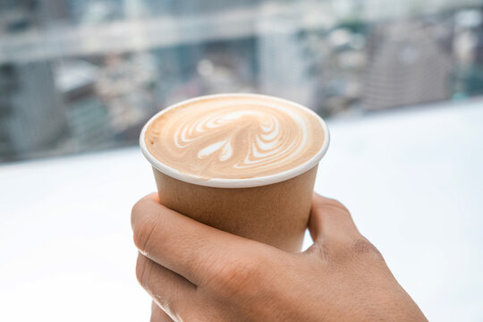 Hand holding paper cup of coffee latte with white foam art against blurred city background in coffee shop. Close up. Shallow depth of field. Healthy drink. City relaxation and modern cafe culture