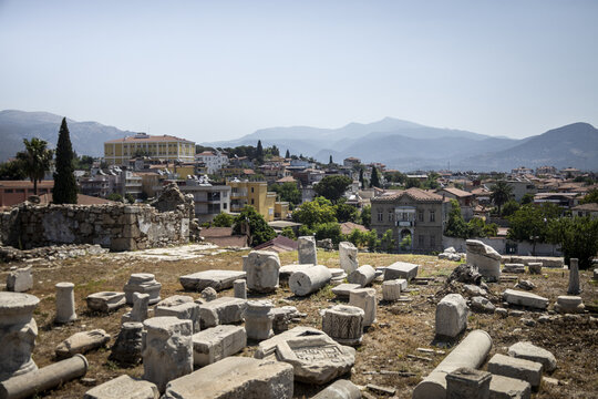 View of ancient marble ruins scattered across a sun-drenched hilltop overlooking a vibrant town nestled against hazy mountains, Milas, Mu&Auml;&Yuml;la, T&Atilde;&frac14;rkiye.