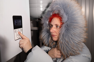 A woman in a warm coat adjusts the thermostat in a chilly environment. She focuses on energy-saving settings to maintain comfort during the cold season while being mindful of resource use.