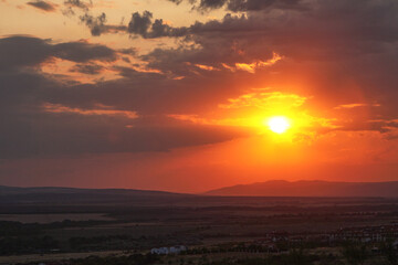 Vibrant sunset over rolling hills with dramatic clouds, casting warm orange and yellow hues across the sky, creating a serene and picturesque landscape at dusk with stunning natural beauty