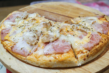 Close-up of a ham, sausage, and cheese Pizza on a wooden tray. Six slices remain (two slices taken), sprinkled with oregano. A popular, cheesy Thai street food snack emphasizing casual dining.