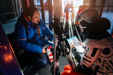 Scenic two skiers study trail map inside ski lift gondola at warm sunrise light at mountain alpine resort. Scenery group friends teamwork, planning, and excitement downhill winter sports adventure