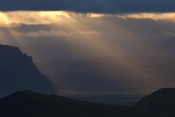 View of celestial rays piercing through a dramatic cloudy sky, illuminating a rugged landscape with contrasting shadows, Dyrholaey, Iceland.