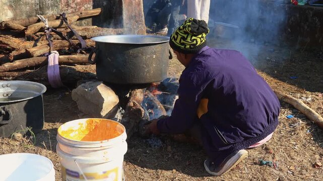 A person cooks outdoors using an open fire. Smoke rises from the flames, with a large pot and various tools set nearby, emphasizing traditional cooking methods. Natural and resourceful setup captured.