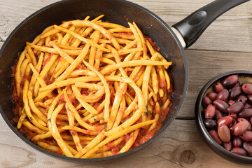 Greek stewed string beans with tomatoes and olive oil (Fasolakia Lathera) in a pan on a rustic wooden table, and a bowl with kalamata olives. Vegan main course or side dish.