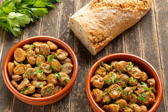 Two clay tapas bowls with a vegan version of Spanish habas con jam&oacute;n on a rustic wooden background. Half a loaf of bread and a bunch of parsley at the back.
