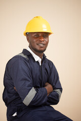 A professional portrait of a confident male African engineer or industrial worker seated with his arms crossed, wearing a navy blue overall and a yellow hard hat.  