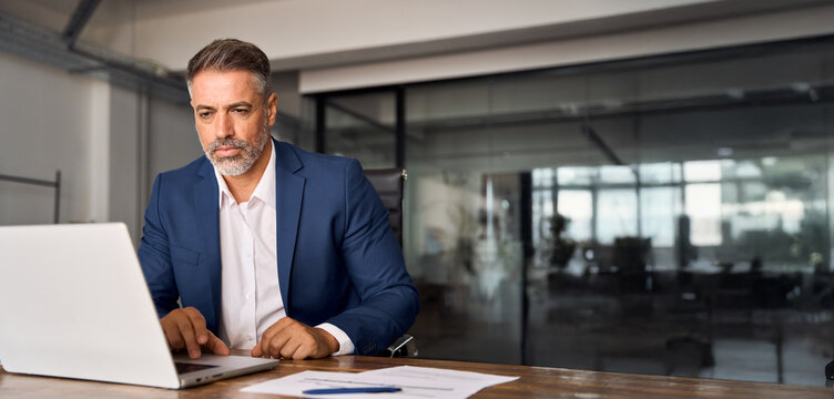 Mature Indian or Latin business man ceo trader using computer, typing, working in modern office doing online data market analysis, thinking planning tech strategy looking at laptop. Banner, copy space
