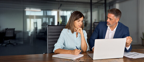 Mature 50s age Latin man mentoring mid age European woman discussing project on laptop pc computer in office. Two colleagues, group of partners professional people working together. Banner, copy space