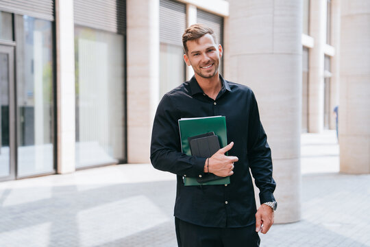 A man in a black shirt smiling, holding folders, standing in a sunlit urban environment.