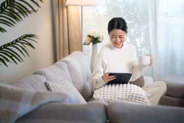 Fototapeta premium Relaxed woman sitting on cozy sofa using digital tablet while holding a cup of coffee.