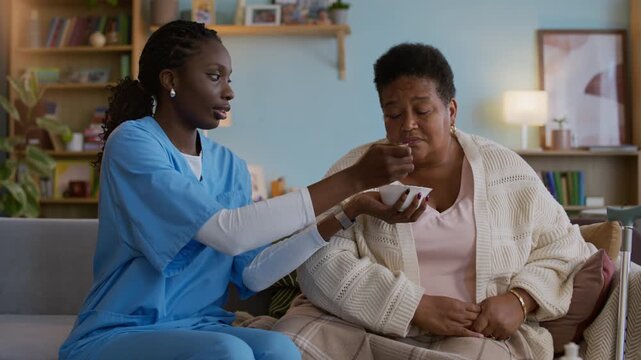 Medium long shot of young African American nurse in medical uniform feeding senior woman with breakfast while visiting at home
