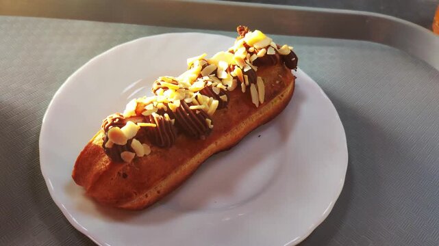 Chocolate Eclair With Almond Flakes On White Plate. Delicious Pastry Dessert Sits On Tray In Sunlight.