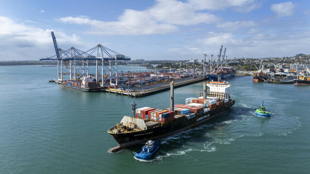 Auckland, New Zealand - 19 September 2025: Aerial view of a container ship, dwarfed by the cranes and a sea of containers at the port, contrasts with the turquoise harbor waters.