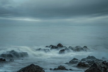 Moody seascape, silky waves flow over rugged rocks with distant headlands under gray winter sky.