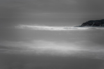 Fototapeta premium Black and white minimalist seascape, smooth water flows past jagged headland with ethereal, misty long-exposure.
