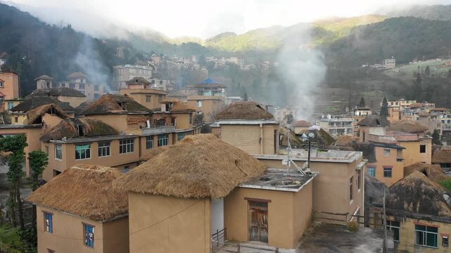 Drone shot over traditional homes with straw rooftops and chimneys billowing smoke in Yuanyang, China
