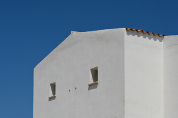 Obraz premium Minimalist white mediterranean-style building with small square windows and a terracotta roof edge, set against a deep blue, cloudless sky.